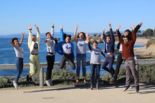 Group of students jumping in the air with the ocean in the background.